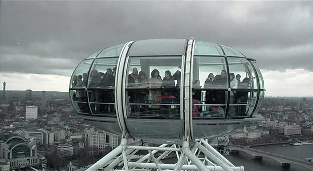 9 Songs (2004) movie still: medium — A glass observation capsule of the London Eye sits high above the urban sprawl. Multiple people stand inside…
