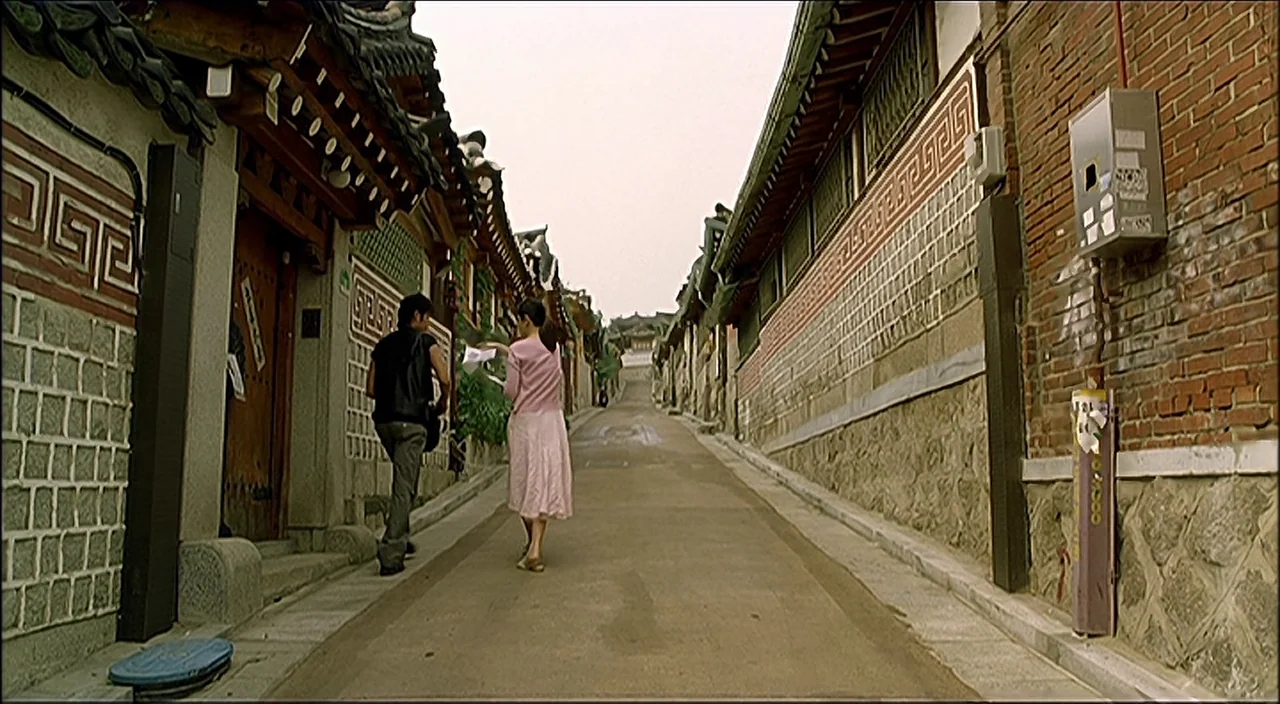 3-Iron (2004) movie still: fullbody — A narrow paved alleyway in a historic district with traditional tiled roofing on walls. Two young adults walk…