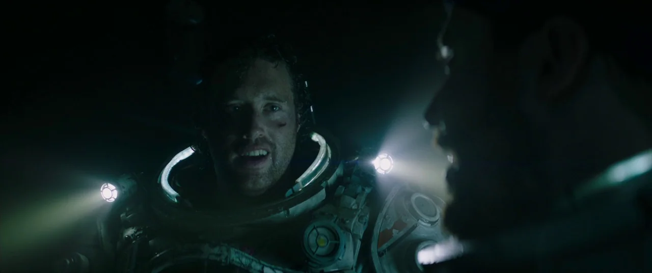 Underwater (2020) movie still: medium — A dark, cramped environment where two people wear bulky mechanical spacesuits with active lights. One man…
