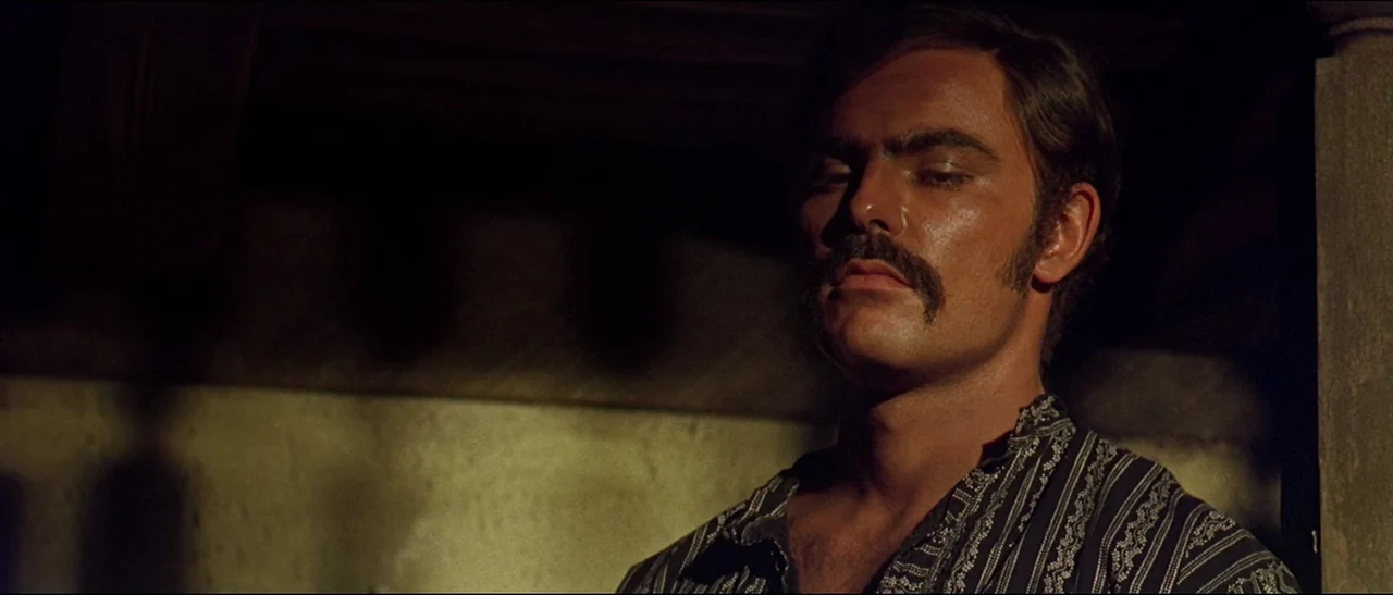 The Appaloosa (1966) movie still: closeup — A dimly lit indoor space with shadows cast across a textured wall. A man with a thick mustache stands…