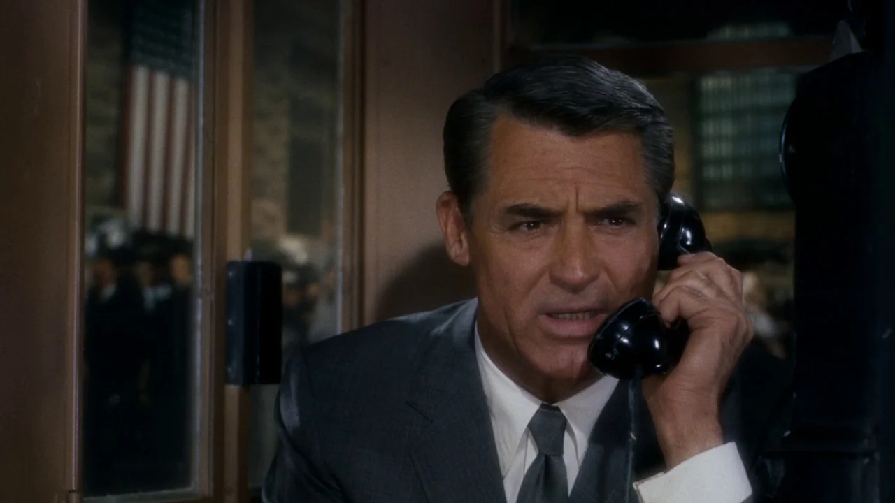 North By Northwest (1959) movie still: closeup — A man stands inside a glass-paned booth located in a large transit hall. The man listens intently…
