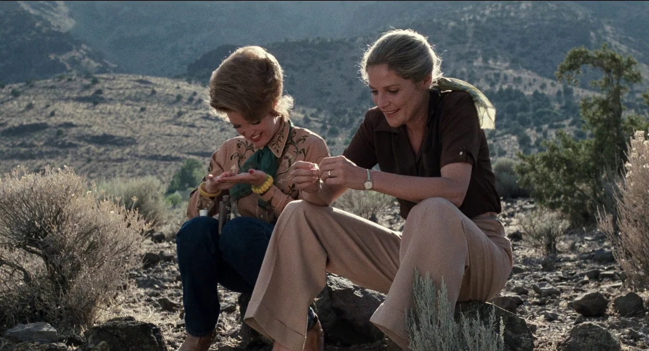 Desert Hearts (1985) movie still: medium — Two women sit on rocks in a dry, sparse desert landscape with mountains behind them. They are focused on…