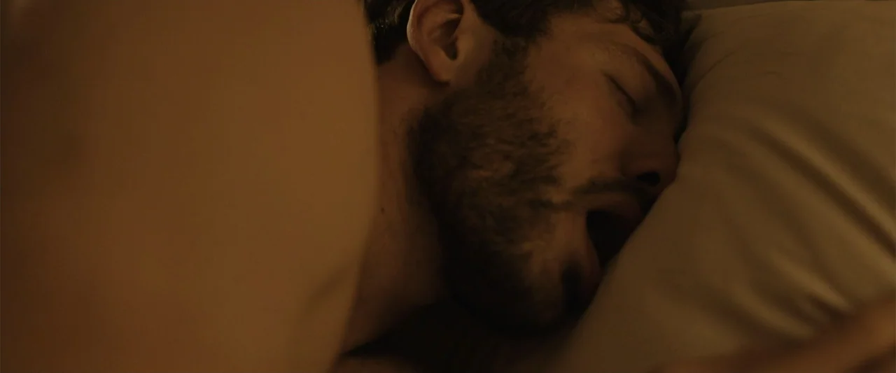 The Thorn On His Side (2014) movie still: closeup — A dark bedroom setting features a person resting in bed on a pillow. A man with short brown hair…