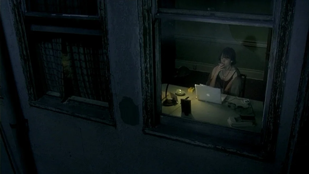 Land of Plenty (2004) movie still: wide — A dark exterior alley view looking through a window into an illuminated workspace. A woman sits at a desk…