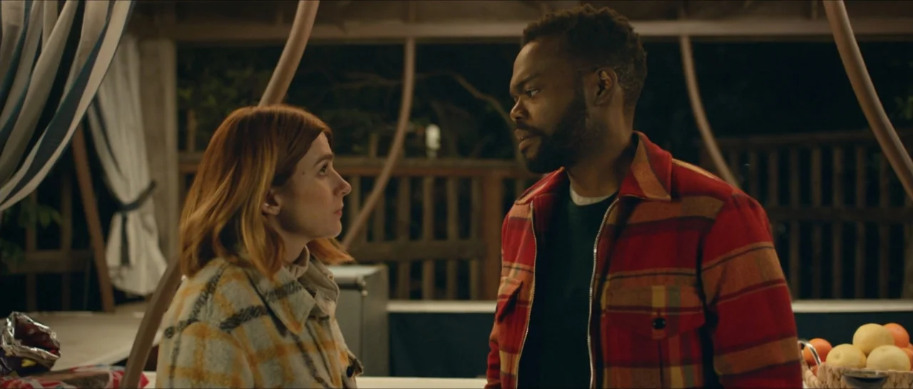We Broke Up (2021) movie still: medium — An outdoor patio area at night with wooden rafters and a striped fabric awning. A man and a woman stand…