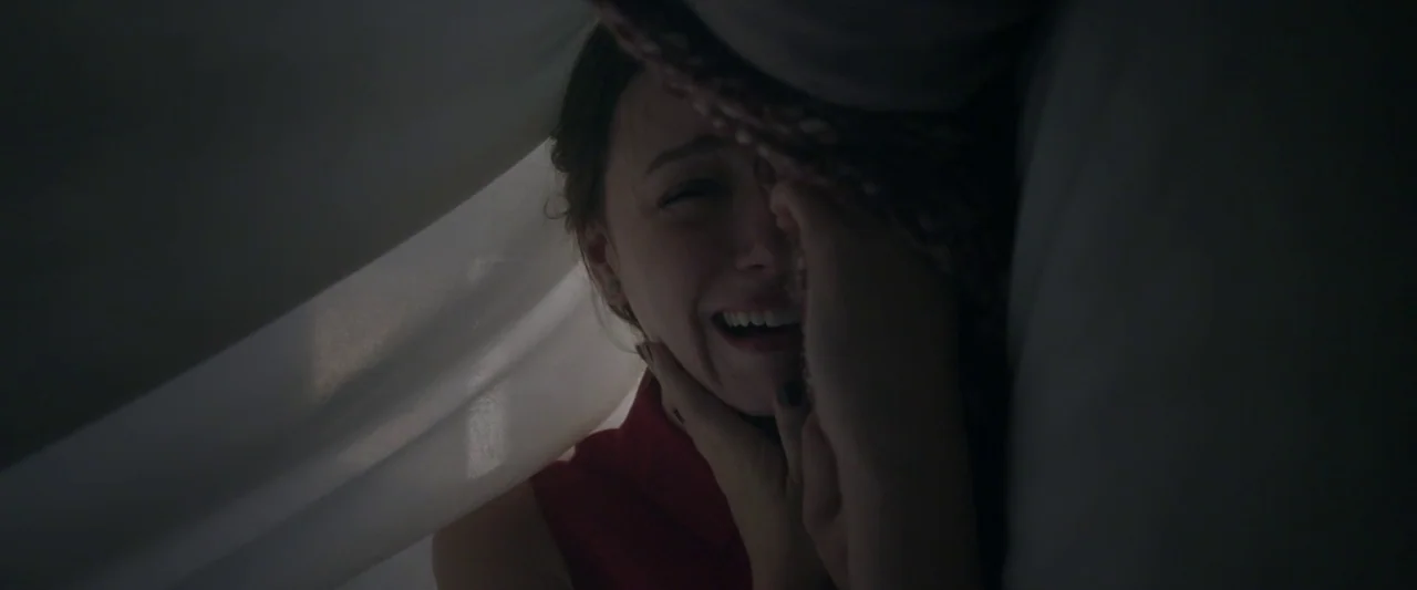 Starry Eyes (2014) movie still: closeup — A dark indoor space where a woman laughs while held under a white curtain. Light filters through the…