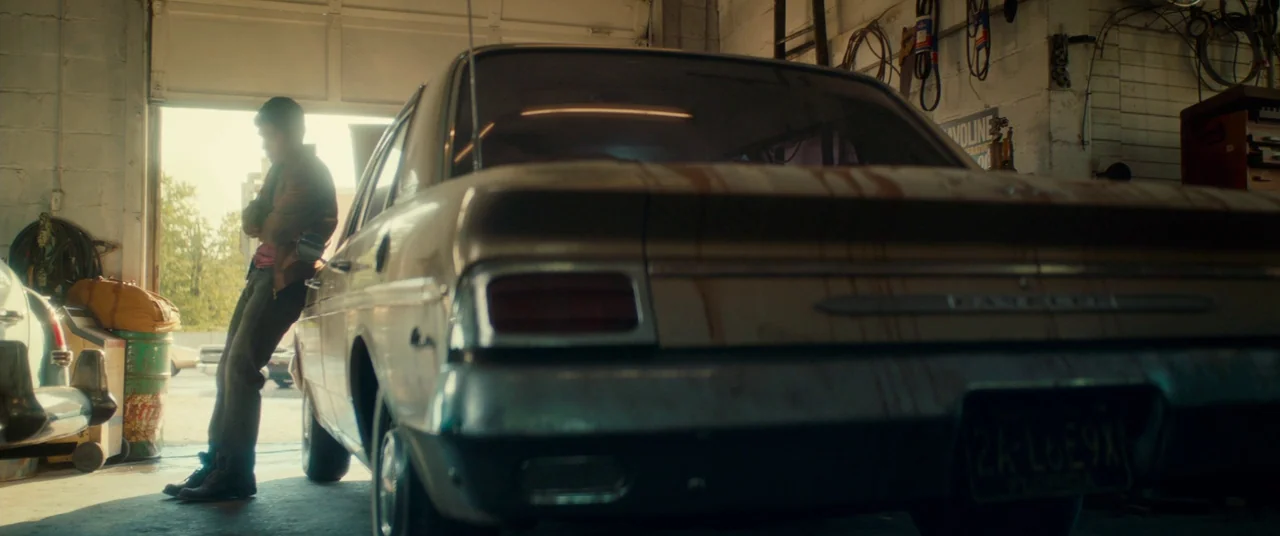 Scary Stories to Tell in the Dark (2019) movie still: fullbody — An auto repair shop features a vintage sedan with rust stains near an open bay. A…