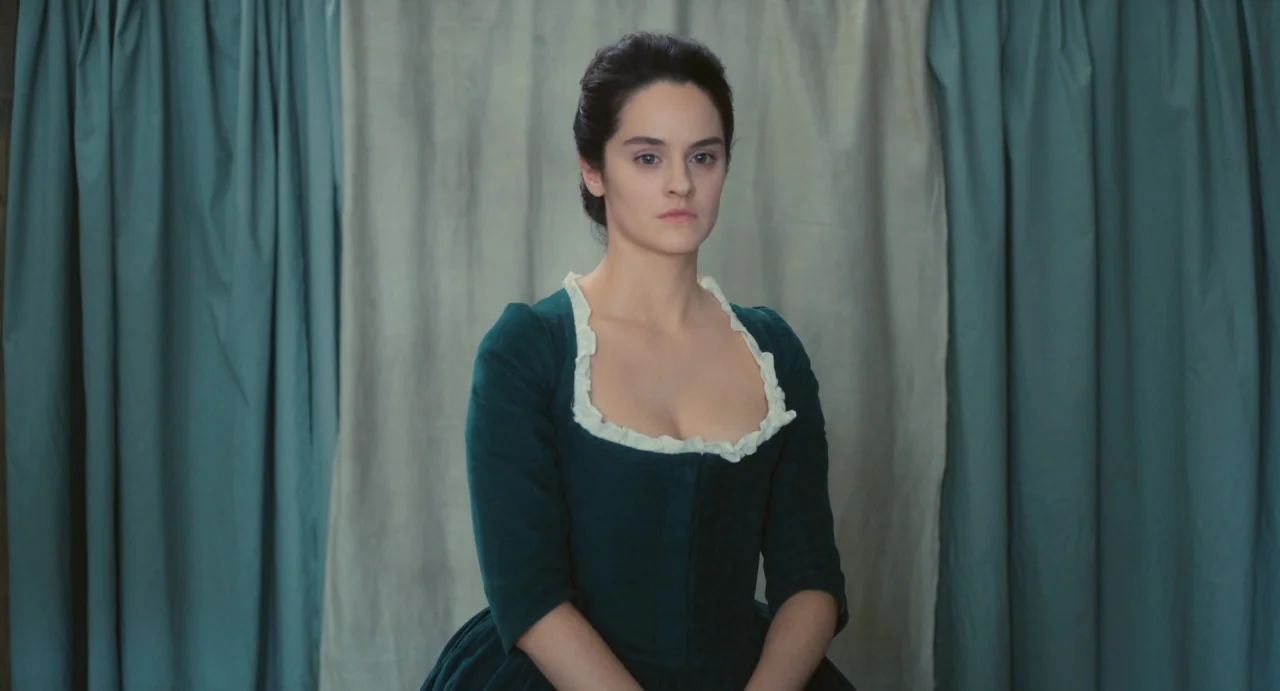 Portrait of a Lady on Fire movie still: medium — A woman stands centered before a backdrop of layered blue-green and beige fabric curtains. She…