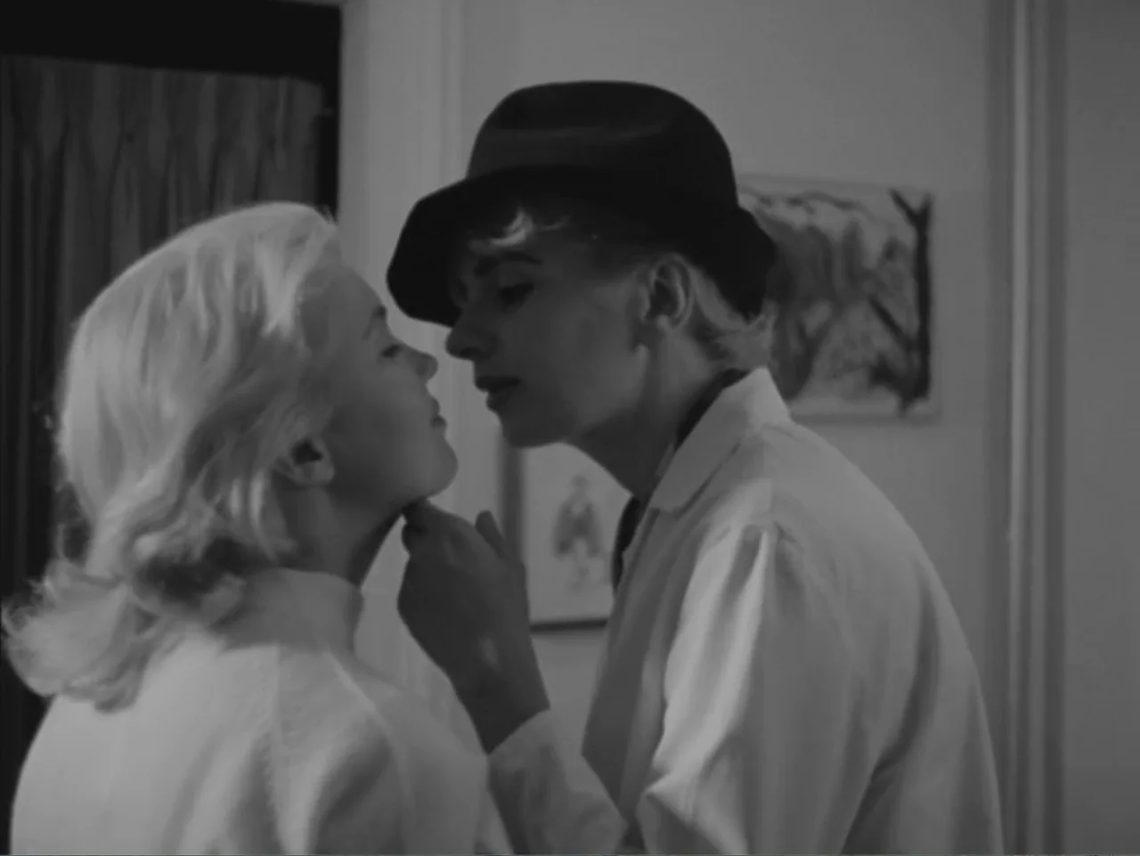 Chained Girls (1965) movie still: medium — A domestic indoor setting with white walls and simple framed art. One woman gently touches the chin of…