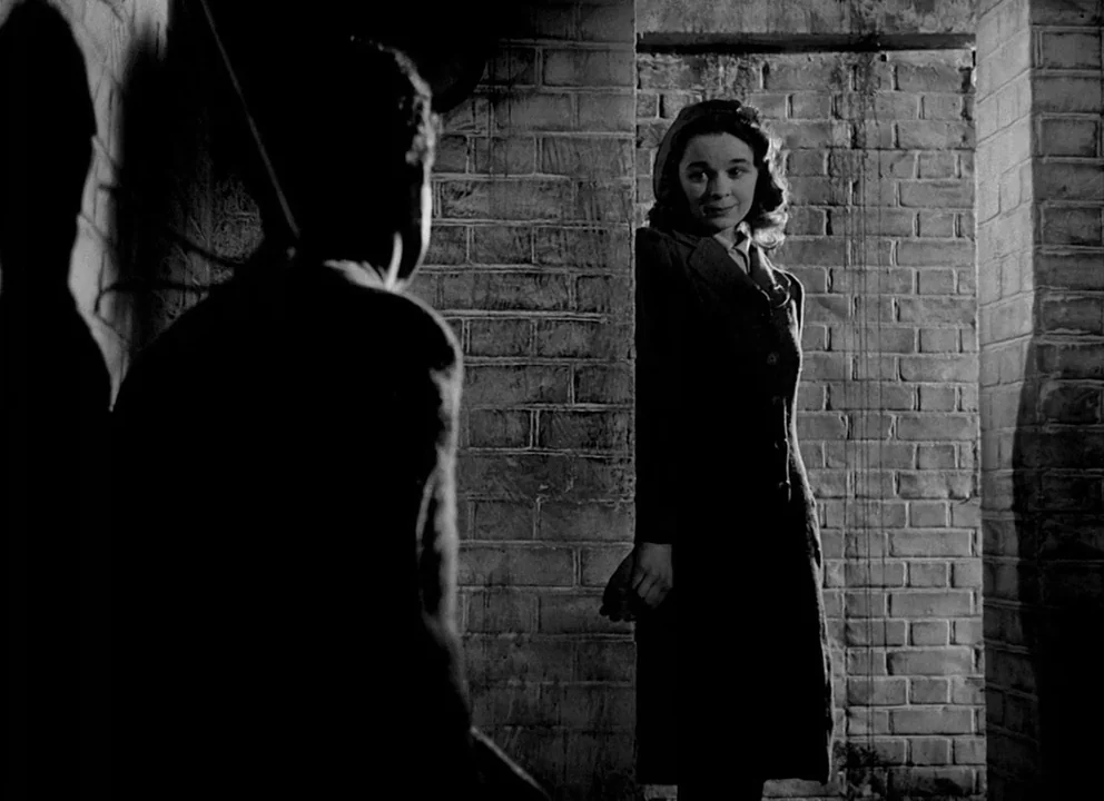 Odd Man Out (1947) movie still: medium — A dark brick-lined corridor with rough mortar joints and heavy shadow contrast. A woman in a dark dress…