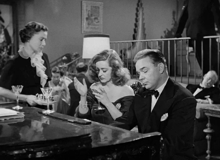 All About Eve (1950) movie still: medium — A refined living room featuring a grand piano and a staircase with an iron railing. A man plays the piano…