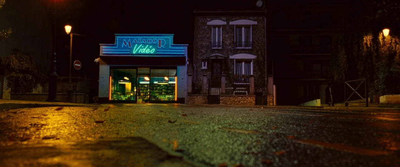 Micmacs (2009) movie still: establishing — A quiet nighttime street features a glowing neon video store beside a stone house. The asphalt road is…