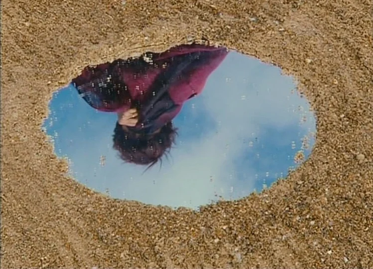 The Beaches of Agnès (2008) movie still: closeup — A circular mirror lies flat on the textured sand of a beach. The mirror reflects a person looking…