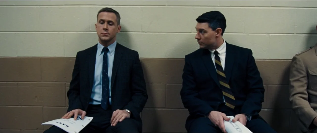 First Man (2018) movie still: medium — A plain waiting area features a split-tone cinder block wall painted beige and tan. Two men in formal…