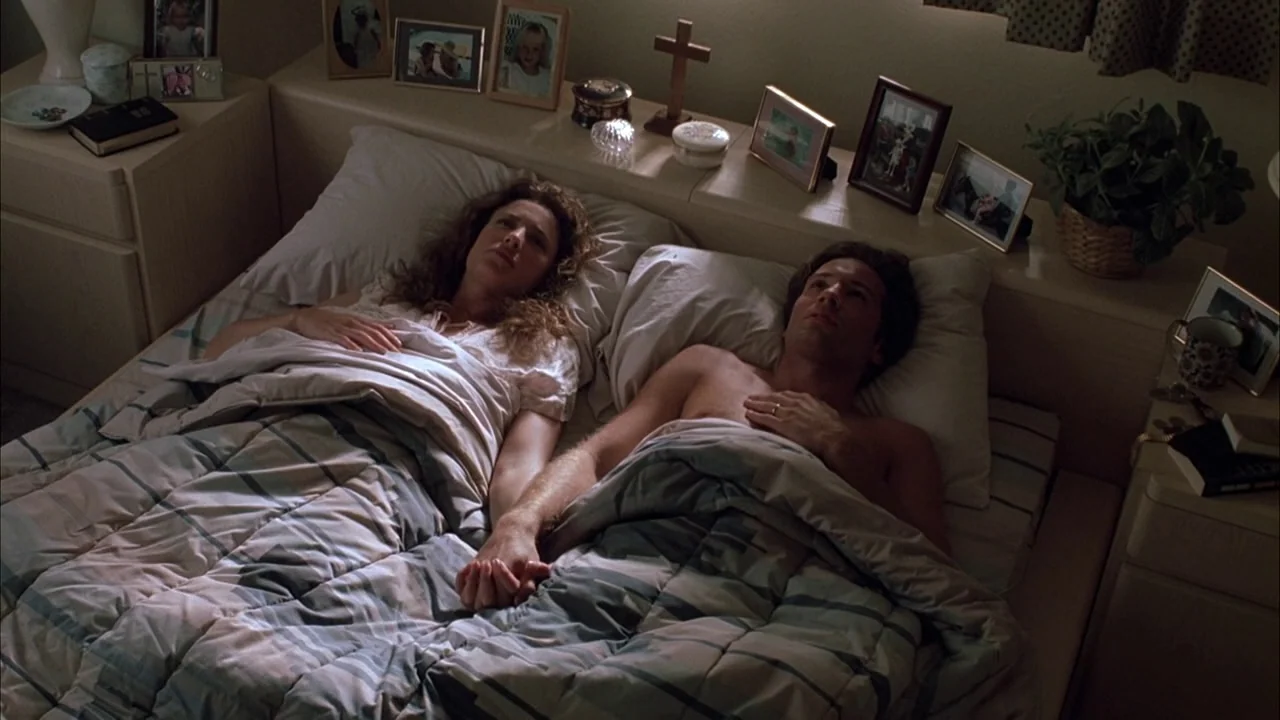 The Rapture (1991) movie still: medium — A bedroom contains a bed with a blue plaid comforter and personal picture frames. A man and a woman lie in…