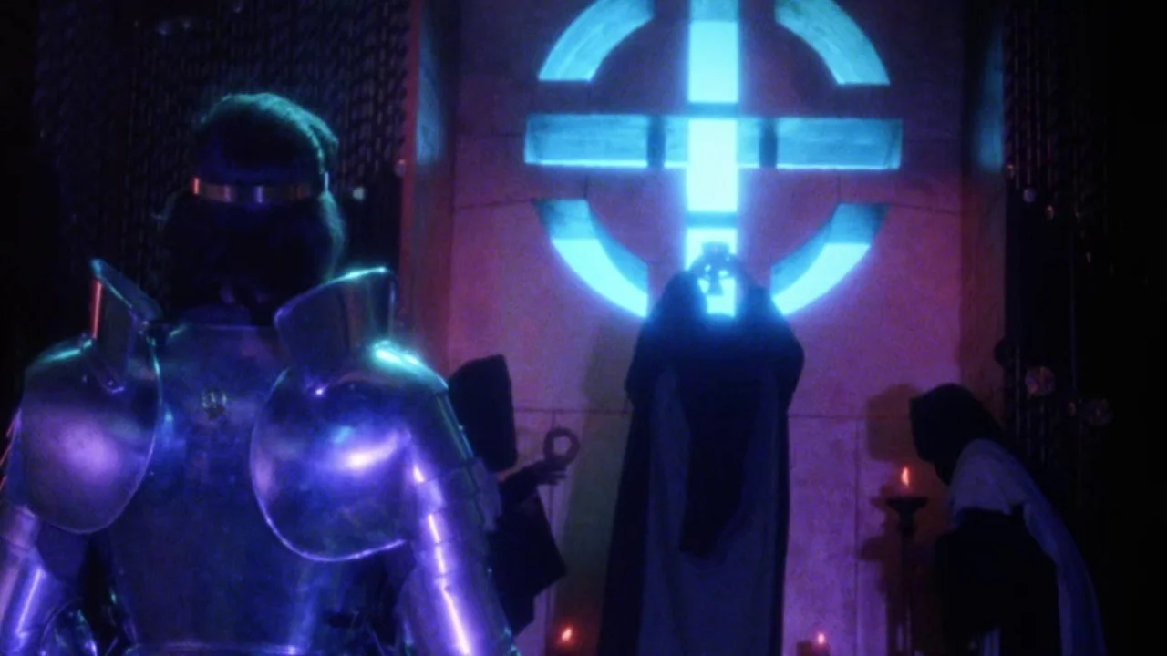 Excalibur (1981) movie still: medium — A stone chamber features a large illuminated cross symbol mounted on the rear wall. A knight in armor stands…