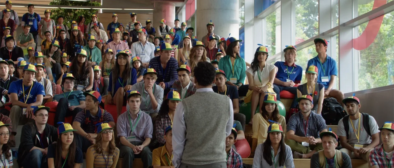 The Internship (2013) movie still: medium — A bright corporate atrium with large floor-to-ceiling windows and tiered seating for attendees. A man in…