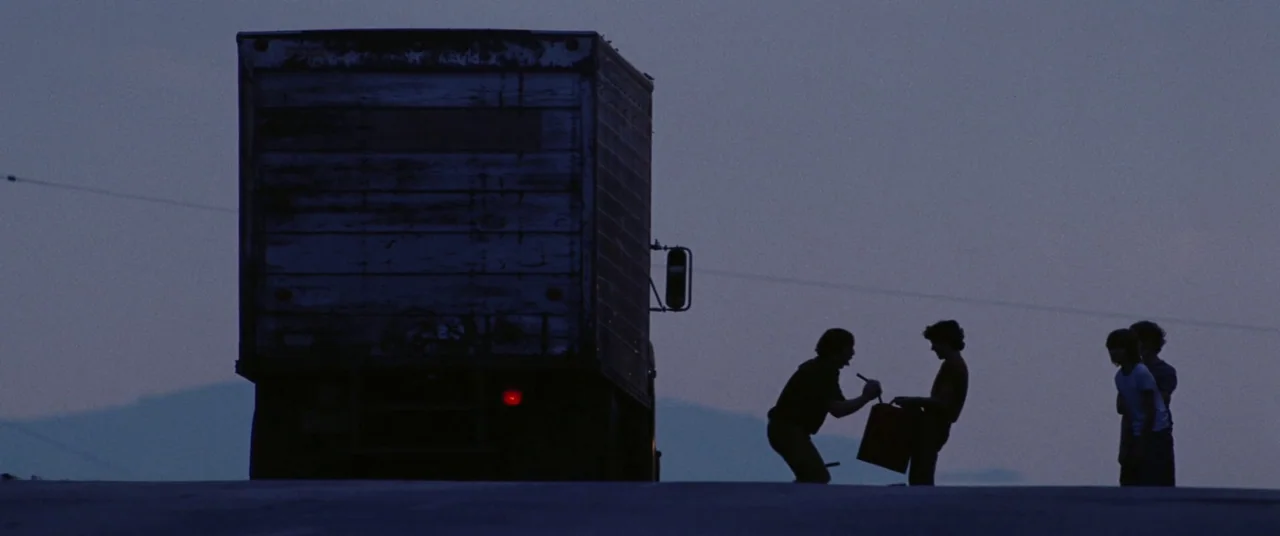 Licorice Pizza (2021) movie still: wide — A box truck sits parked on the shoulder of a desolate paved road at dusk. Several teenagers stand in…