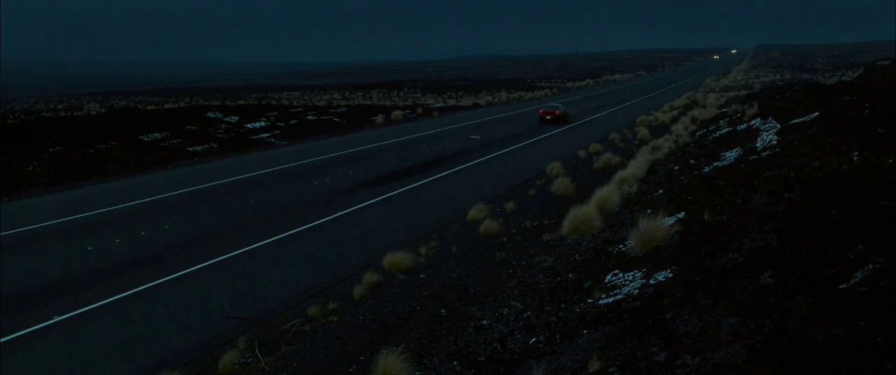 The Descendants (2011) movie still: wide — A long paved road cuts through a desolate, dark volcanic landscape under a night sky. A lone car moves…