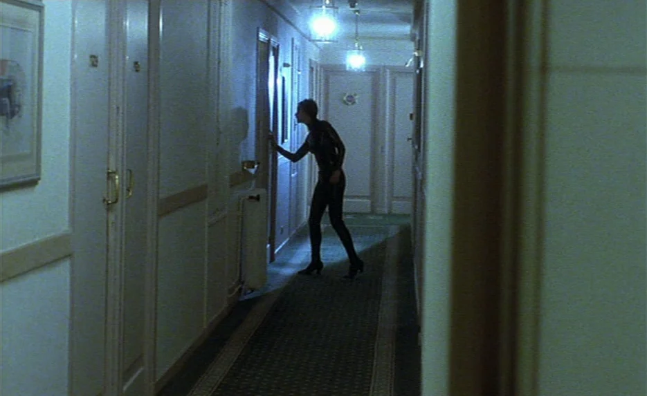 Irma Vep (1996) movie still: fullbody — A long, dimly lit hotel corridor features closed doors and a patterned carpet runner. A woman in a dark…