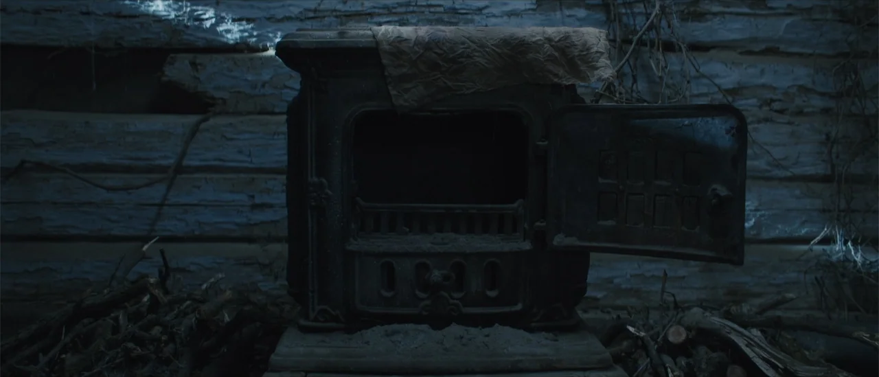 All Fun and Games (2023) movie still: medium — A dark, rustic interior features an antique cast-iron wood stove against a log wall. The stove door…