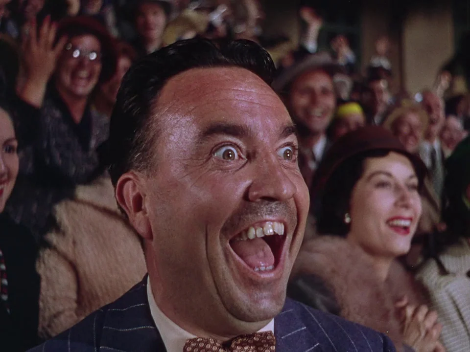 Singin’ in the Rain (1952) movie still: closeup — A crowded public event with spectators visible in the blurred background. A middle-aged man shouts…