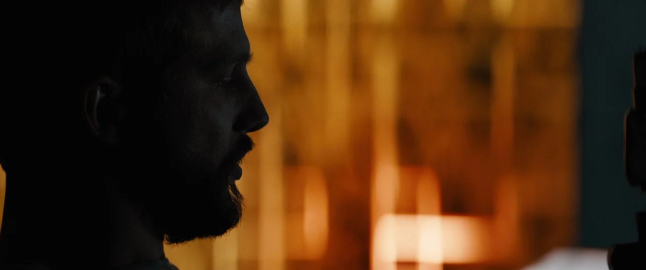 Upgrade (2018) movie still: closeup — A dimly lit interior space with a blurred, warm-toned light source in the background. A man in profile stands…