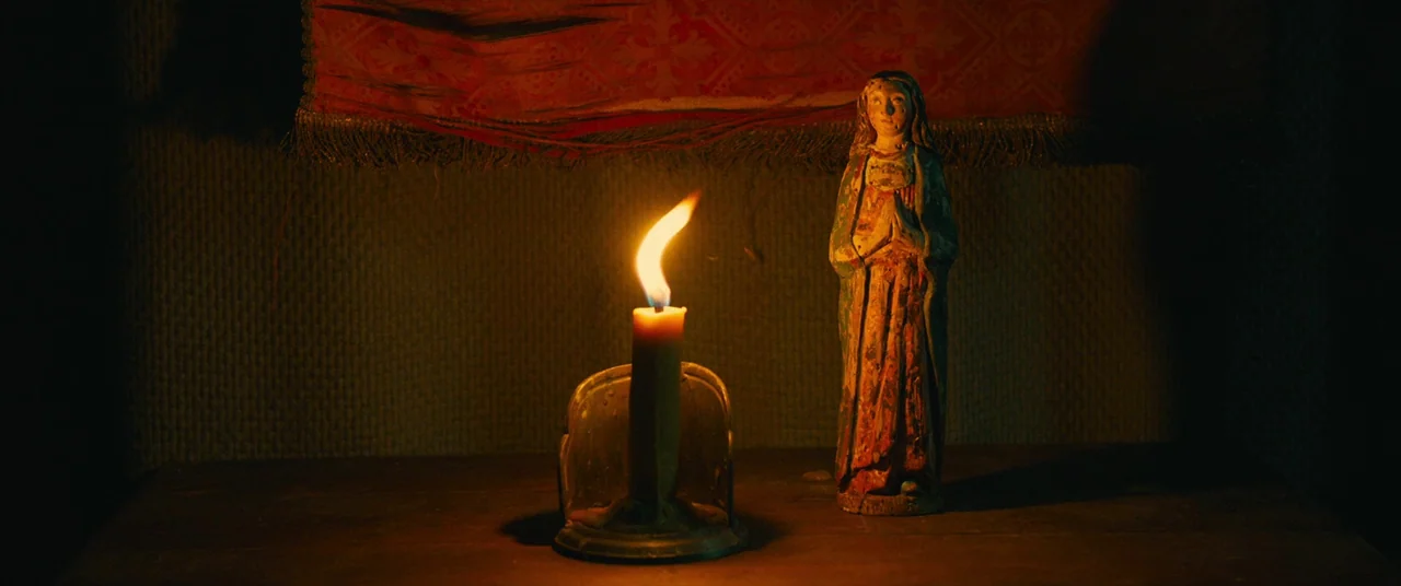 Benedetta (2021) movie still: medium — A small wooden statue of a religious figure stands on a dark wooden surface. A lit candle sits in a metal…