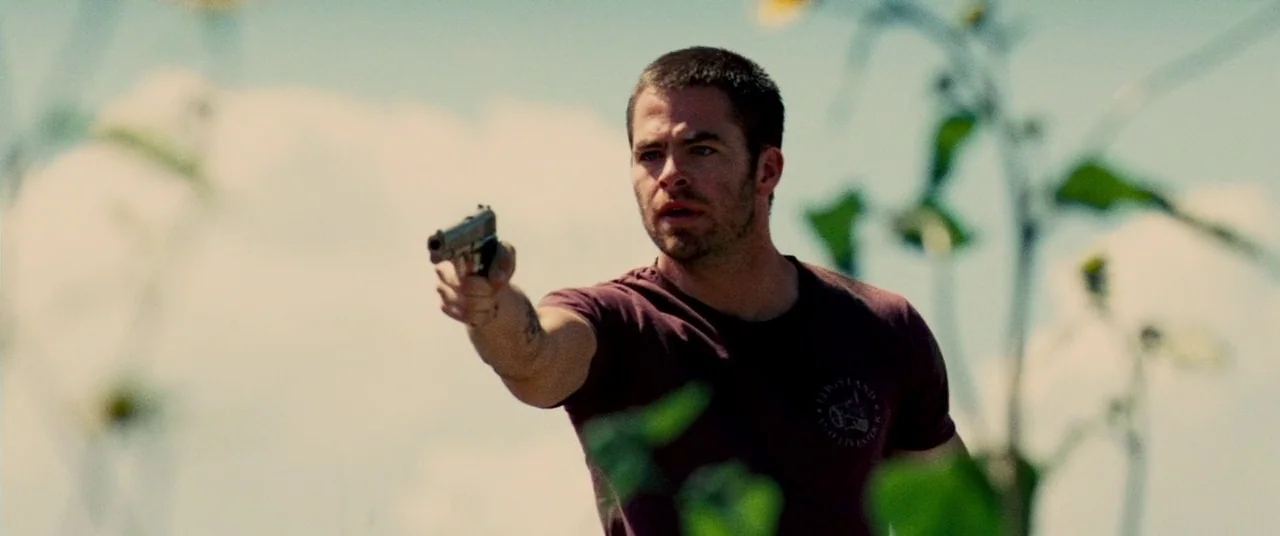 Carriers (2009) movie still: medium — A man stands in an outdoor field with tall weeds and blurred sunflowers. He holds a pistol outstretched toward…