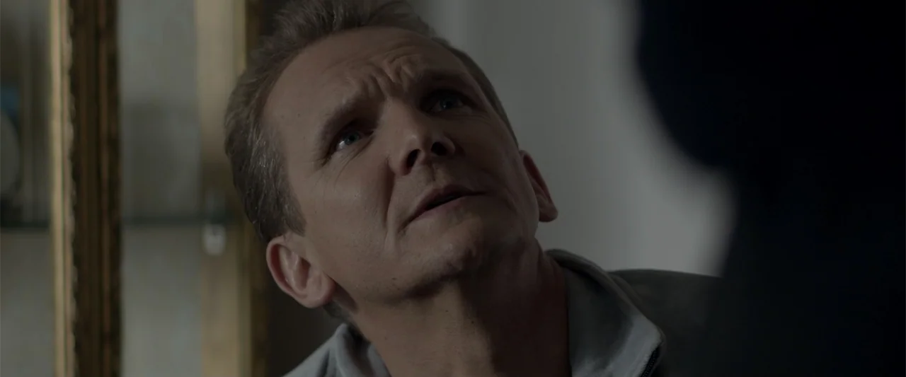 A Walk Among the Tombstones (2014) movie still: closeup — A dimly lit indoor space with neutral walls and a blurred foreground silhouette present. A…