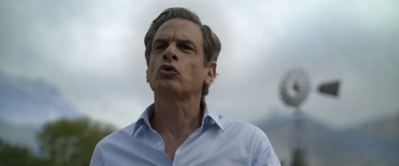 Doublespeak (2021) movie still: medium — A man stands outdoors in an open rural field under a heavy grey sky. The man in a blue dress shirt speaks…