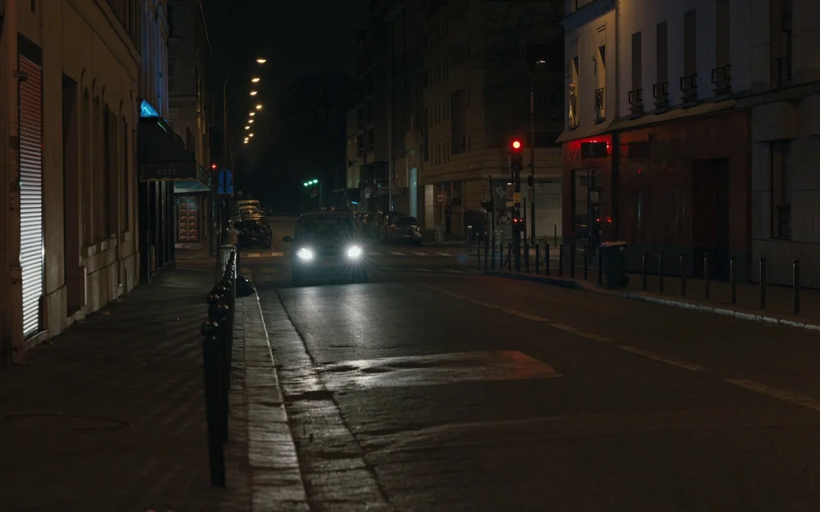 Let The Sunshine In (2017) movie still: wide — A quiet urban street at night with multi-story buildings lining the dark asphalt road. A single…