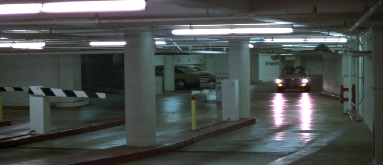 The Long Goodbye (1973) movie still: wide — A concrete parking garage interior features white support pillars and a striped entry barrier. A car…