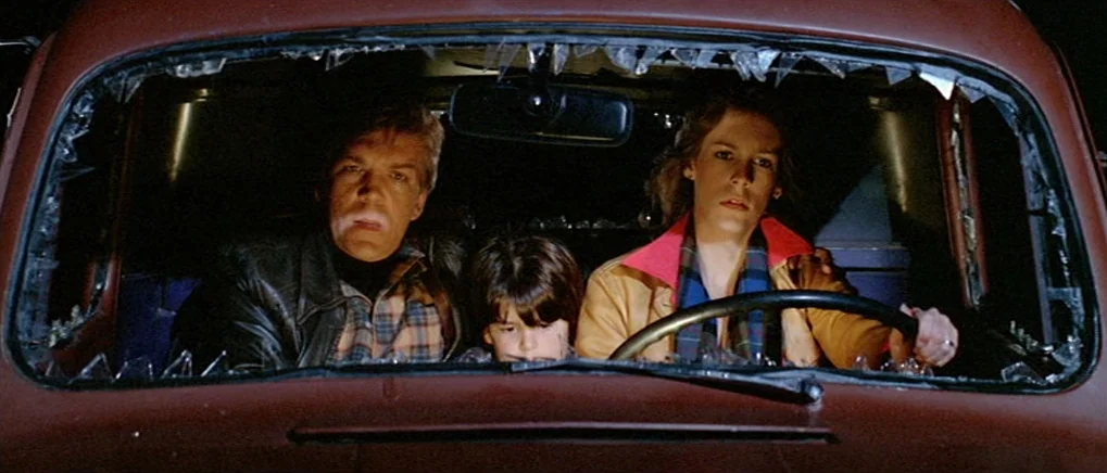 The Fog (1980) movie still: medium — A dark, cramped car cabin features a shattered windshield with jagged glass shards remaining. A man, a young…
