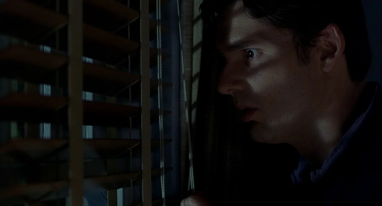 Hulk (2003) movie still: closeup — A man peers through a set of horizontal window blinds in a dark room. Deep shadows hide parts of his face as…