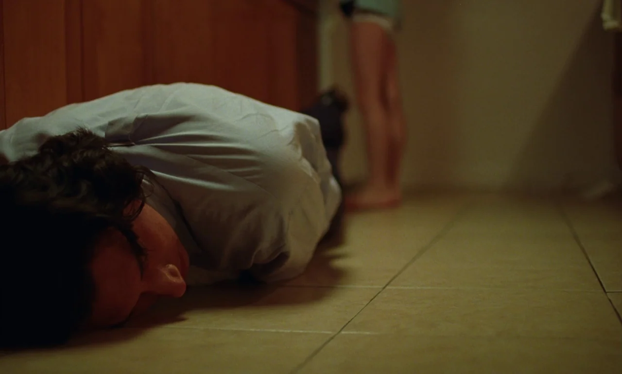 Marriage Story movie still: medium — A man lies on a beige tile floor in a dimly lit home. A second person stands upright nearby in the background…