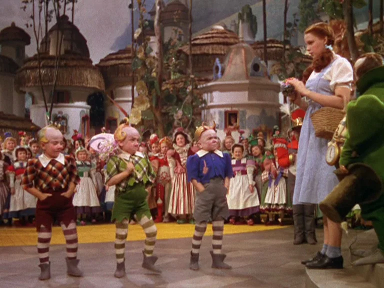 The Wizard Of Oz (1939) movie still: fullbody — A whimsical fantasy village features small thatched-roof houses and a yellow path. Three small men…