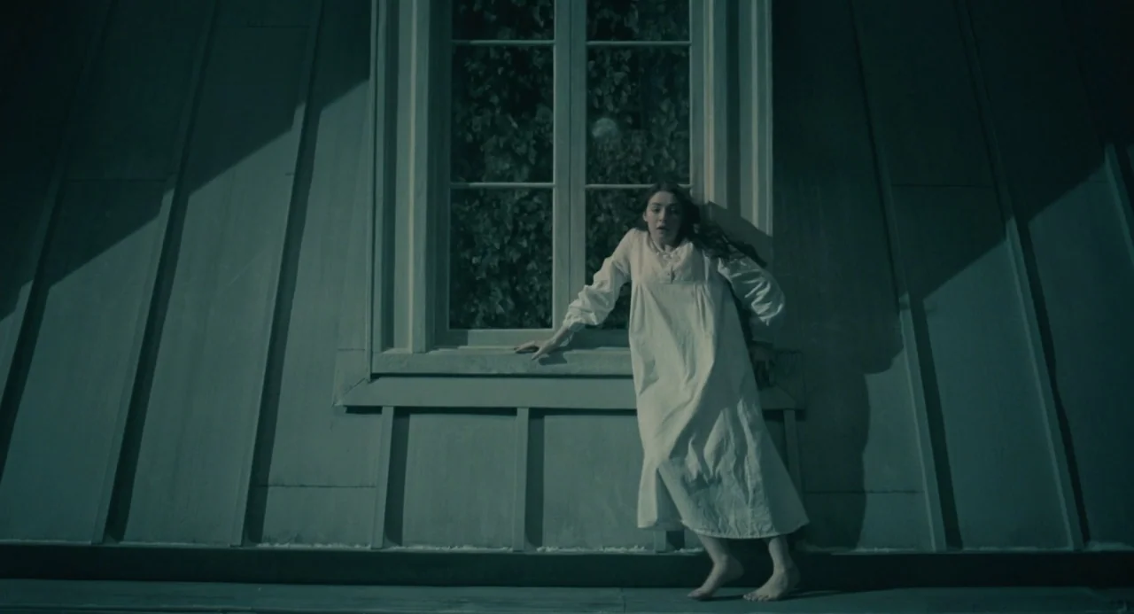 The Moth Diaries (2011) movie still: fullbody — A woman in a white nightgown stands on a steep, dark metal roof. She leans against the exterior…