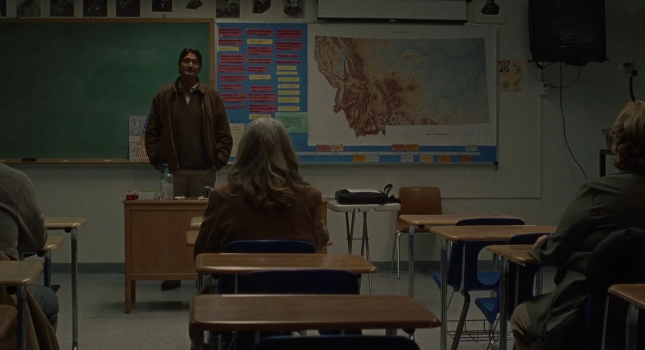 Certain Women (2016) movie still: wide — A school classroom featuring a green chalkboard, a large wall map of Montana, and desks. A man in a brown…