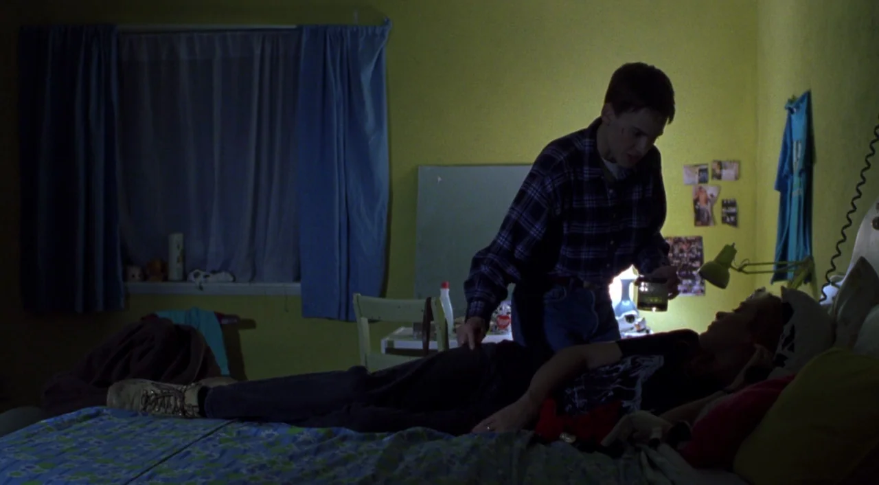 Boys Don’t Cry (1999) movie still: medium — A dimly lit bedroom features mustard-yellow walls and a blue-curtained window. A young man stands over a…