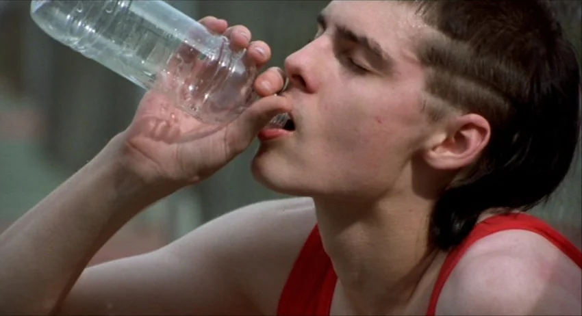 Gummo (1997) movie still: closeup — A teenage boy in a red tank top stands on a sports field. He tilts his head back while drinking from a clear…