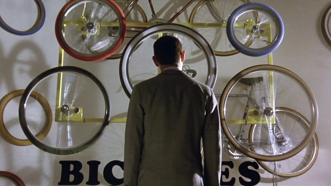 Pee-Wee’s Big Adventure (1985) movie still: medium — A man stands before a cream-colored wall decorated with various mounted bicycle wheels. The…