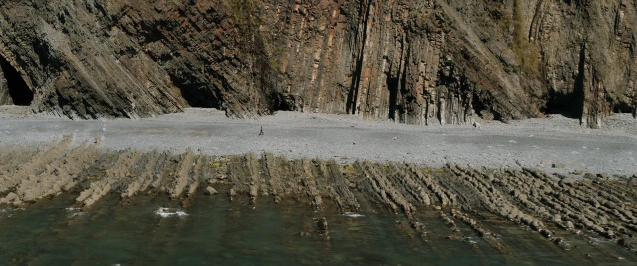 Rebecca movie still: wide — A desolate, jagged coastline features stratified cliff faces rising above a narrow pebble beach. A solitary person walks…
