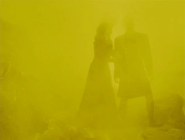 Careful (1992) movie still: fullbody — A rocky, barren landscape is completely saturated in a thick, opaque yellow fog. Two human figures stand in…