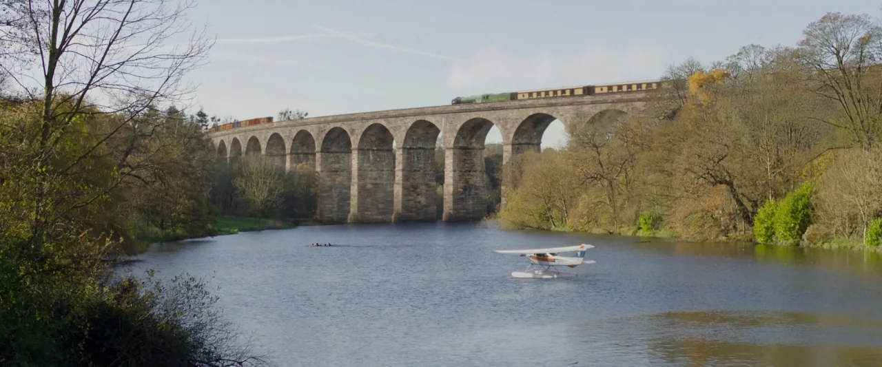 Paddington 2 (2017) movie still: wide — A stone multi-arch viaduct spans a wide river flanked by dense, thinning autumn trees. A vintage steam train…