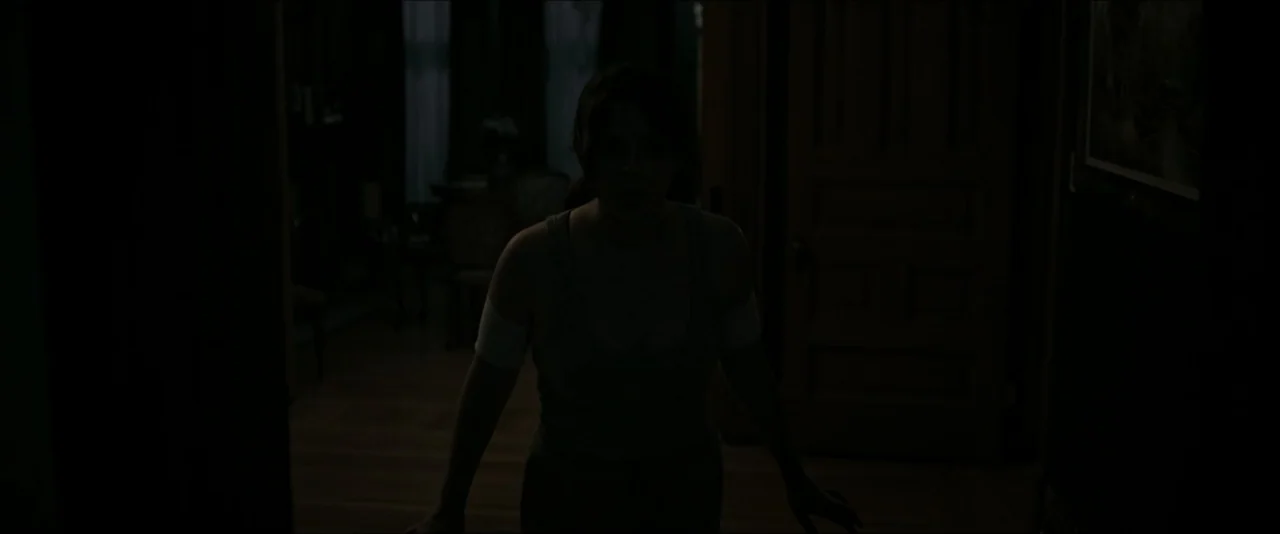 It Feeds (2025) movie still: medium — A dark, residential interior hallway with a visible doorway leading to an adjoining room. A young woman stands…