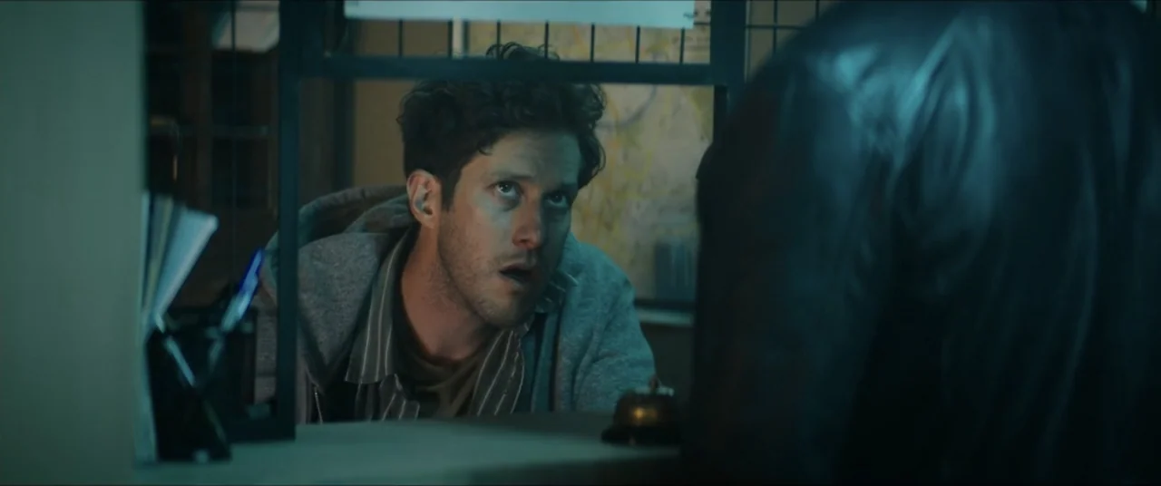 Man with No Past (2025) movie still: medium — A dark, partitioned service area with a metal service counter and a bell. A young man in a hooded…