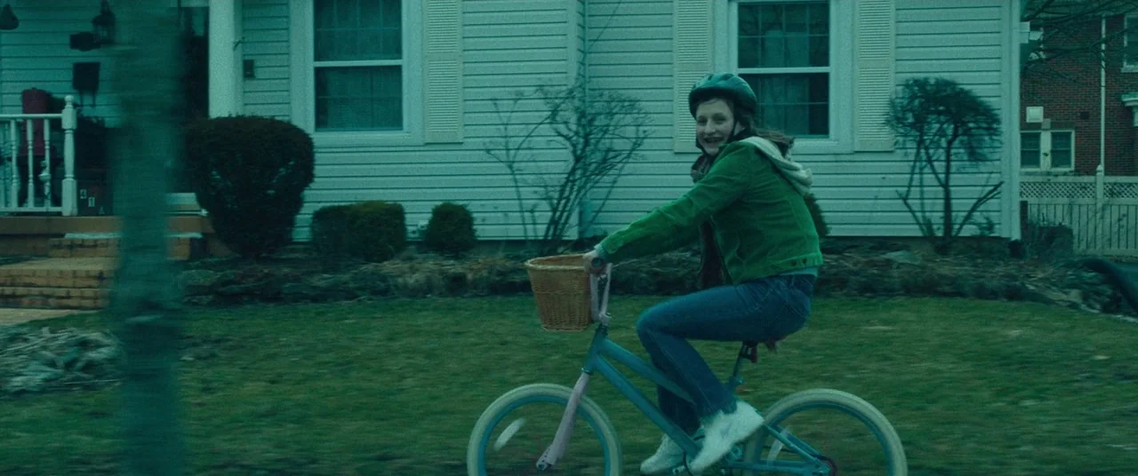 Dark Waters (2019) movie still: fullbody — A suburban house with white siding stands behind a patch of grass. A girl rides a light blue bicycle with…