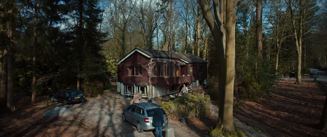 Speak No Evil (2022) movie still: wide — A two-story dark brown wooden house sits nestled in a dense, deciduous forest. Three people stand near a…