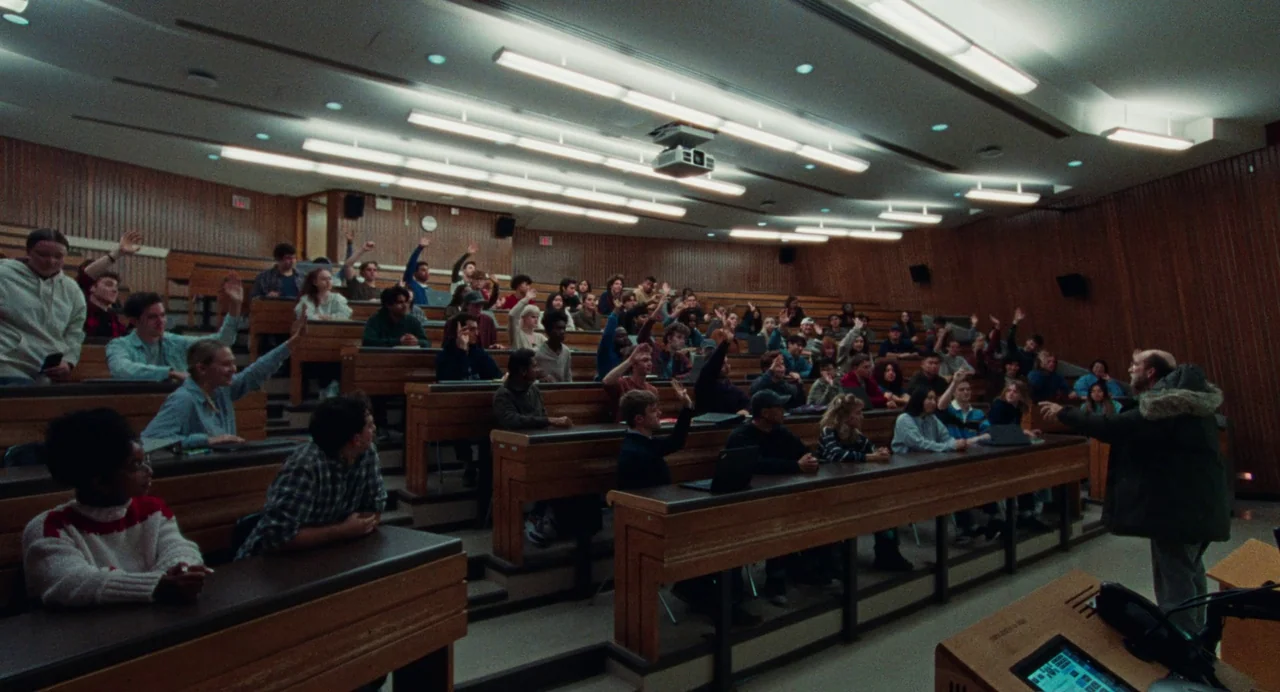Dream Scenario (2023) movie still: wide — A tiered university lecture hall features rows of wooden desks and panelled wood walls. Numerous students…